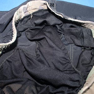 May include: A pair of black swim trunks featuring an interior black mesh liner and an elasticized waistband with a camouflage pattern in shades of tan, brown, and green. The garment includes a drawstring closure and visible stitching details.