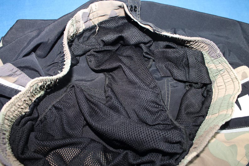 May include: A pair of black swim trunks featuring an interior black mesh liner and an elasticized waistband with a camouflage pattern in shades of tan, brown, and green. The garment includes a drawstring closure and visible stitching details.