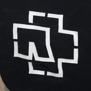 May include: A black fabric garment featuring a large white stylized cross logo printed on the front. To the left, a vertical panel displays a multi-colored camouflage pattern in shades of brown, green, and tan, separated by a thin white line.
