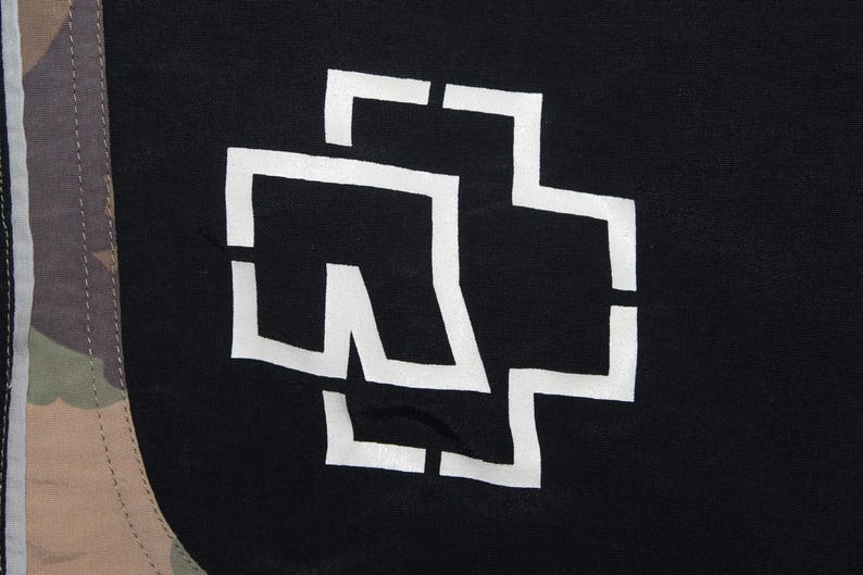 May include: A black fabric garment featuring a large white stylized cross logo printed on the front. To the left, a vertical panel displays a multi-colored camouflage pattern in shades of brown, green, and tan, separated by a thin white line.