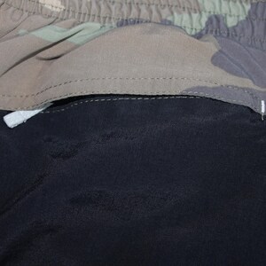 May include: A pair of shorts featuring a solid black section and a camouflage patterned section in shades of green, brown, and tan. The garment includes an elasticized waistband and a horizontal seam separating the two distinct fabric panels.
