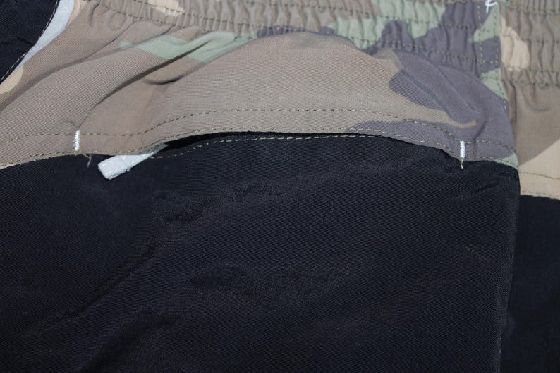 May include: A pair of shorts featuring a solid black section and a camouflage patterned section in shades of green, brown, and tan. The garment includes an elasticized waistband and a horizontal seam separating the two distinct fabric panels.