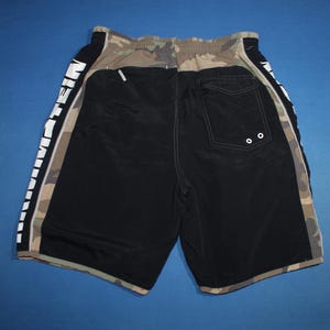 May include: Black and camouflage patterned swim shorts featuring an elastic waistband and a back pocket with two white grommets. The side panels display the word EINSTEIN in large white capital letters running vertically along the leg.