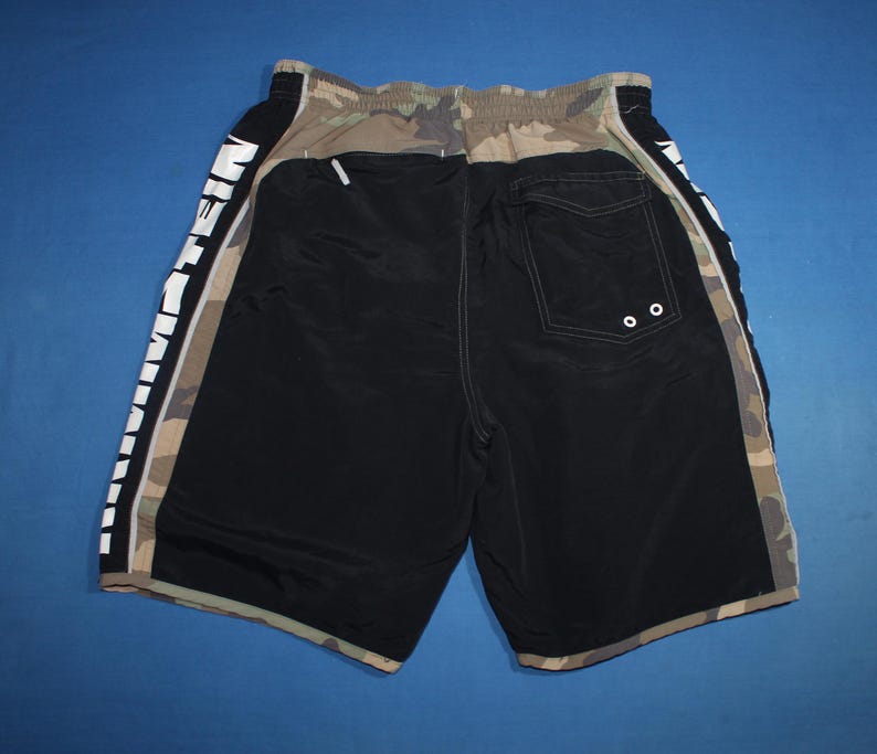 May include: Black and camouflage patterned swim shorts featuring an elastic waistband and a back pocket with two white grommets. The side panels display the word EINSTEIN in large white capital letters running vertically along the leg.