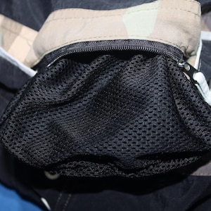 May include: A black garment featuring a zippered pocket with a black mesh lining. The fabric surrounding the pocket displays a tan and brown camouflage pattern, while the main body of the item is solid black with white lettering visible on the side.