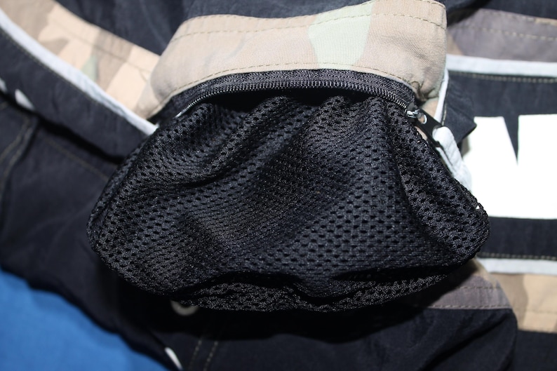 May include: A black garment featuring a zippered pocket with a black mesh lining. The fabric surrounding the pocket displays a tan and brown camouflage pattern, while the main body of the item is solid black with white lettering visible on the side.