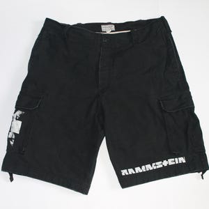 May include: Black cargo shorts featuring a white Rammstein logo printed on the lower left leg and a white graphic design on the right cargo pocket. The shorts include a button closure, belt loops, and two side cargo pockets with flap closures.
