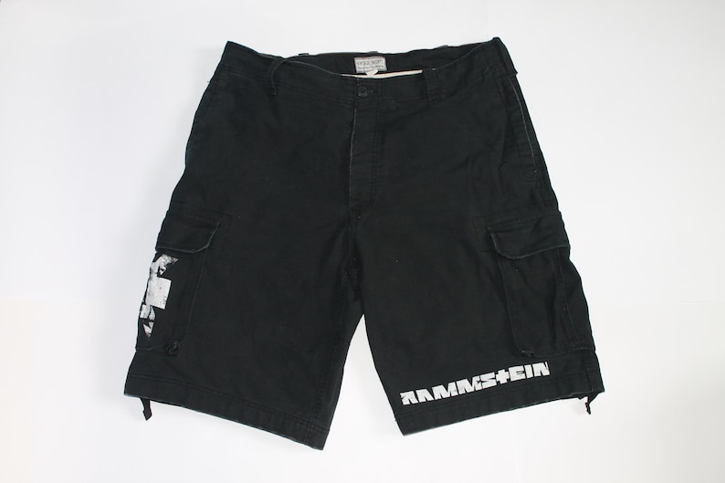 May include: Black cargo shorts featuring a white Rammstein logo printed on the lower left leg and a white graphic design on the right cargo pocket. The shorts include a button closure, belt loops, and two side cargo pockets with flap closures.