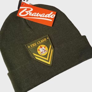 May include: Olive green knit beanie with a patch that reads "THE CLASH" and a Bravado tag. The patch is olive green with yellow and white accents. The beanie is a classic style, perfect for cold weather.