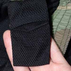 May include: A pair of shorts featuring a green, brown, and black camouflage pattern on the exterior waistband and a black mesh fabric lining on the interior. The garment includes a rectangular black mesh pocket attached to the inner lining.