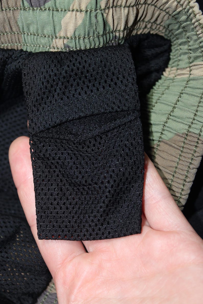 May include: A pair of shorts featuring a green, brown, and black camouflage pattern on the exterior waistband and a black mesh fabric lining on the interior. The garment includes a rectangular black mesh pocket attached to the inner lining.