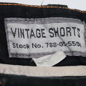 May include: A black garment featuring a rectangular grey fabric label with the text VINTAGE SHORTS and Stock No. 788-05-559. Below the label is a smaller white tag partially visible with the text 100% C. The waistband has orange stitching.