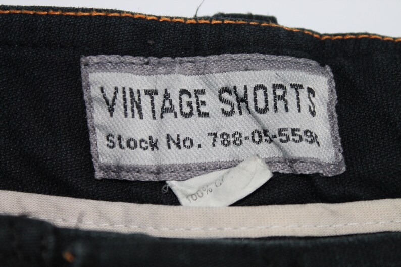 May include: A black garment featuring a rectangular grey fabric label with the text VINTAGE SHORTS and Stock No. 788-05-559. Below the label is a smaller white tag partially visible with the text 100% C. The waistband has orange stitching.
