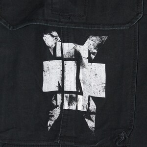 May include: A black fabric garment featuring a large, distressed white graphic print of a stylized cross symbol. The design is centered on a rectangular pocket flap with visible stitching and a horizontal seam across the top section of the fabric.