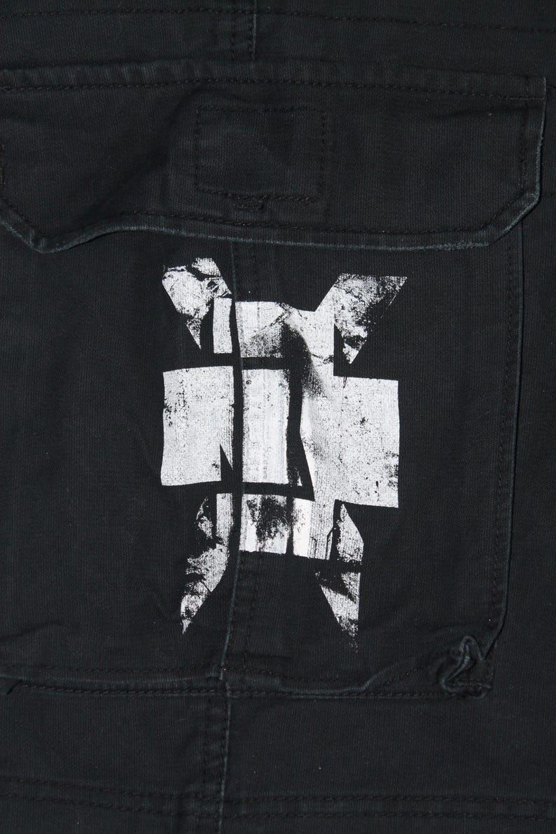 May include: A black fabric garment featuring a large, distressed white graphic print of a stylized cross symbol. The design is centered on a rectangular pocket flap with visible stitching and a horizontal seam across the top section of the fabric.