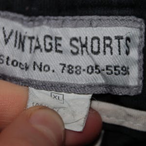 May include: A black garment featuring a rectangular grey fabric label with black text that reads VINTAGE SHORTS and Stock No. 788-05-559. Below this is a smaller white tag displaying the size XL and partially visible text indicating 100% cotton.