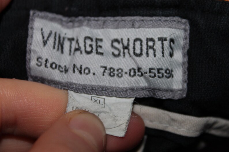 May include: A black garment featuring a rectangular grey fabric label with black text that reads VINTAGE SHORTS and Stock No. 788-05-559. Below this is a smaller white tag displaying the size XL and partially visible text indicating 100% cotton.