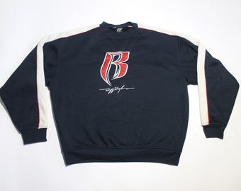 Op de afbeelding: Marineblauw sweatshirt met een rood en wit logo en handtekening. De top met lange mouwen heeft witte strepen over de mouwen en een ronde hals. Het sweatshirt is een casual stijl.