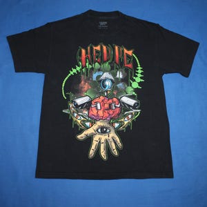 May include: Black t-shirt with a graphic design featuring the word "HELIE" in green and orange, along with a surreal illustration of a brain, eye, and hand. The design includes green and white accents.