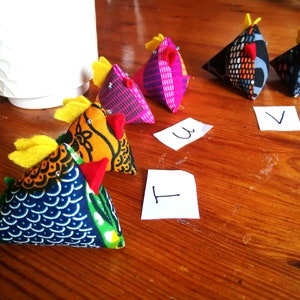 May include: A collection of colorful fabric chicken ornaments with yellow crests and red wattles. The chickens are made from various patterned fabrics, including blue, orange, and pink designs. Small white cards with numbers are scattered around the chickens.