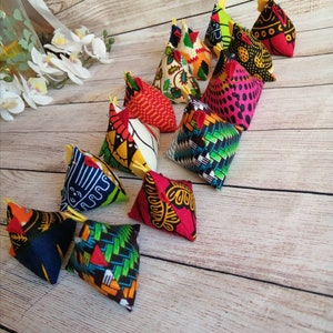 May include: A collection of 12 colourful fabric chicken-shaped pincushions. Each pincushion is a different pattern and colour, including red, yellow, blue, green, and black. The pincushions are arranged in a row on a wooden surface.