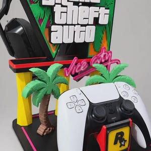 Op de afbeelding: Een zwarte, gele en roze gaming displaystandaard met het Grand Theft Auto Vice City logo, twee groene palmbomen en een Rockstar Games embleem. De standaard houdt een wit-zwarte gamecontroller en een zwarte headset op de achterste steun.