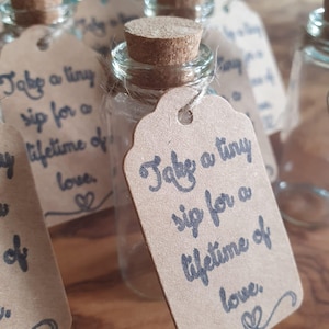 May include: A collection of small glass bottles with cork stoppers and brown tags. Each tag is inscribed with the phrase "Take a tiny sip for a lifetime of love" in dark blue script, with a heart symbol.