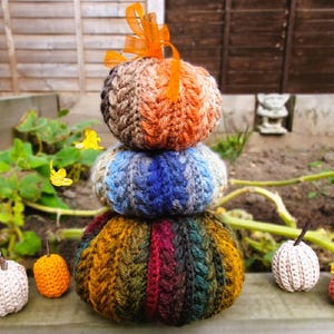 May include: A trio of crocheted pumpkins in autumnal colours: brown, blue, and green, with an orange ribbon. Smaller crocheted pumpkins in orange, white, and pink are arranged nearby, creating a seasonal display. The pumpkins are made of yarn.