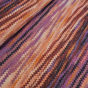Könnte beinhalten: Ein Stoffballen mit einem sich wiederholenden Zickzackmuster in den Farben Violett, Orange, Dunkelrot und Schwarz. Das Textil weist ein sichtbares metallisches Schimmern im gesamten Gewebe auf, wobei die Zickzacklinien diagonal über die Oberfläche verlaufen.