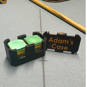 May include: A black pool cue chalk holder containing two green chalk cubes. A black lid with the text "Adam's Case" in orange is next to the holder. A pool cue is visible in the background. The chalk cubes have yellow and black labels.