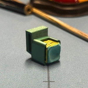 Può includere: Un piccolo oggetto rettangolare verde con un quadrato color oro su un lato. L'oggetto ha una forma squadrata e sembra essere fatto di metallo. Un bastoncino di legno e altri oggetti sono sullo sfondo.