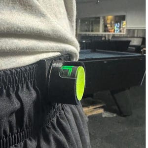以下が含まれることがあります： 黒いレベルツールが黒いスポーツパンツに取り付けられています。 レベルには明るい緑色の気泡とネオンの緑色のエンドキャップが付いています。 その人は白いスウェットシャツを着ています。