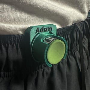 Könnte beinhalten: Ein grünes 3D-gedrucktes Zubehörteil, das an schwarzen Shorts befestigt ist. Das Zubehör trägt den Namen "Adam" und hält einen zylindrischen Gegenstand mit grünem Schaumstoff außen. Der Gegenstand ist teilweise sichtbar.