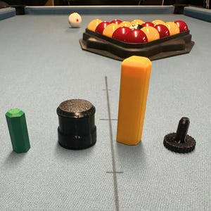 May include: A pool table with a rack of billiard balls, a cue ball, and several accessories. The accessories include a green chalk holder, a black cue tip shaper, a yellow cue shaft protector, and a black cue tip shaper.