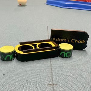 Peut inclure: Un porte-craie de billard noir et jaune avec deux craies et un support. Les craies sont vertes et jaunes. Le texte "Adam's Chalk" est sur un morceau de bois noir.