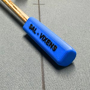 May include: A blue pool cue handle with the text "SAL. VIXENS" in black. The handle is attached to a wooden cue stick. The cue is resting on a grey surface with a cross and a line.
