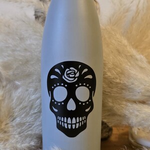 Op de afbeelding: Een grijze waterfles van roestvrij staal met een zwart-wit suiker schedelontwerp. De schedel heeft een bloem op zijn voorhoofd.