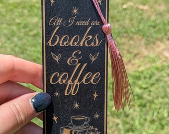 Può includere: Segnalibro nero con la scritta incisa "All I need are books & coffee". Il segnalibro ha un nappa rosa e un disegno di libri, fiori e una tazza di caffè. Lo sfondo è verde sfocato.