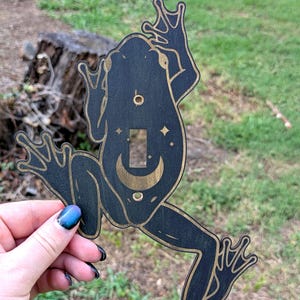 May include: Black wooden frog-shaped light switch cover with gold accents. The frog has a crescent moon and star design. The cover is held up against a background of green grass and a tree stump.