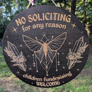 Op de afbeelding: Een rond zwart houten bord met goudkleurige gegraveerde tekst die NO SOLICITING for any reason en children fundraising WELCOME leest. Het ontwerp bevat een centrale mot, twee kristallen met bloemaccenten en kleine sterren, opgehangen aan een touwtje.