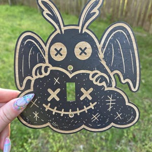 Könnte beinhalten: Schwarze und beige Holz-Lichtschalterabdeckung mit einer cartoonartigen Kreatur mit durchgestrichenen Augen und einem genähten Lächeln, vor einer Wolke. Das Design umfasst Flügel und einen hasenartigen Kopf.