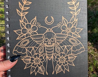 Può includere: Un taccuino a spirale nero con un'illustrazione a linee color oro sulla copertina. Il disegno raffigura una falena con il corpo a forma di teschio, circondata da elementi floreali e da un simbolo di luna crescente in alto.
