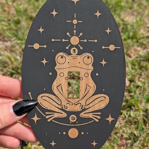 Puede incluir: Cubierta de interruptor de luz ovalada de madera negra con un diseño de rana cortado con láser. La rana está centrada, rodeada de símbolos celestiales y estrellas. La rana y los símbolos son de color marrón claro, contrastando con el fondo negro.
