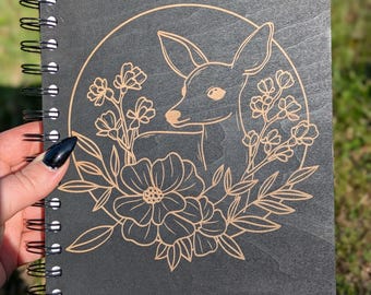 Può includere: Un taccuino nero a spirale con un design di cervo e fiori. Il cervo e i fiori sono incisi in un colore marrone chiaro, su uno sfondo grigio scuro. Il taccuino è tenuto in mano da una persona.