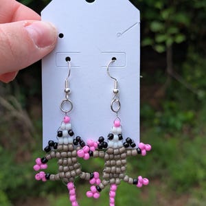 May include: A pair of handmade beaded earrings featuring opossum designs. The opossums are crafted with brown, white, black, and pink beads, with silver-tone ear wires. The earrings are displayed on a white earring card against a blurred green background.