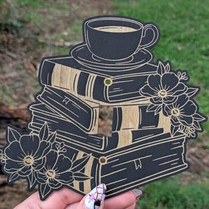 May include: Black and gold wooden wall decor featuring a stack of books with a coffee cup on top. The design includes floral accents and a cutout for a light switch. A decorative piece for a reading nook.