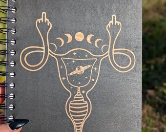 Può includere: Un taccuino a spirale con copertina grigio scuro. La copertina presenta un'illustrazione dorata di un utero con un design celeste, tra cui pianeti, stelle e lune crescenti. Il design incorpora due mani che fanno il dito medio.