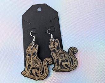 May include: A pair of wooden earrings featuring a skeleton cat design. The earrings are black with a brown wood grain finish and have silver hooks.