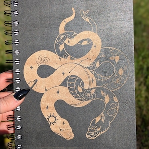 Pu&ograve; includere: Un taccuino a spirale con copertina grigio scuro. La copertina presenta una dettagliata illustrazione di due serpenti intrecciati di colore marrone chiaro, adornati da elementi celesti e floreali. Il taccuino &egrave; tenuto da una mano con un'unghia nera.