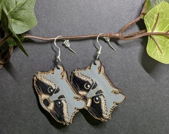 May include: A pair of raccoon-shaped earrings. Each earring features a gray face with black accents around the eyes and a light brown nose. The earrings are made of wood and have silver-colored hooks.